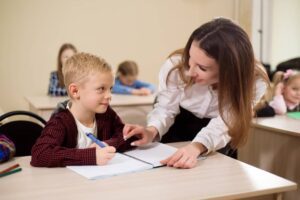 Primary school teacher helping a girl writing at her desk.