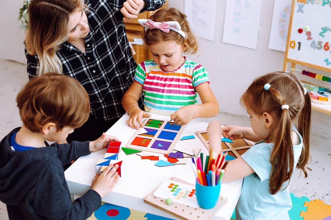 Teacher sitting at table with colored pencils, board games with pupils children playing.