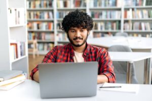 Male student, sitting at table with laptop in university library, working on a project.