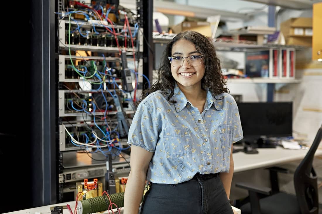 Female student standing in electrical engineering lab and smiling at camera.