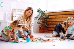 Young female teacher sitting near colorful pencils, different toys with children, checking looking at painting on red paper in bright classroom.