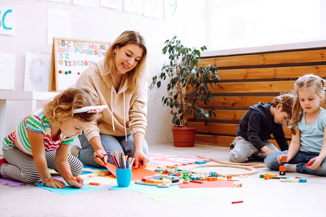 Young female teacher sitting near colorful pencils, different toys with children, checking looking at painting on red paper in bright classroom.